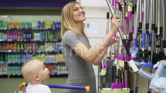 年轻漂亮的妈妈正在超市里挑选拖把而她的小孩则坐在购物车里视频的预览图