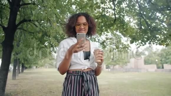 一位美国黑人女士在城市公园里用智能手机走路喝咖啡视频的预览图