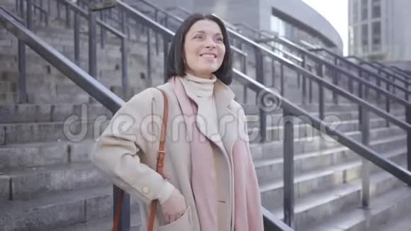 积极的白种人女人站在楼梯上穿着优雅的外套挥动着邀请某人的手阳性的深色头发视频的预览图