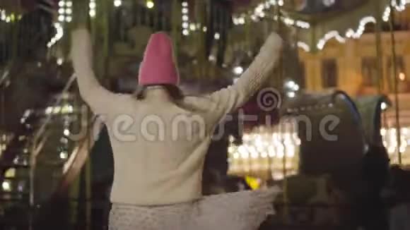 开朗的白种人女孩戴着粉红色的帽子和白色的毛衣在户外五颜六色的旋转木马前旋转积极青年视频的预览图