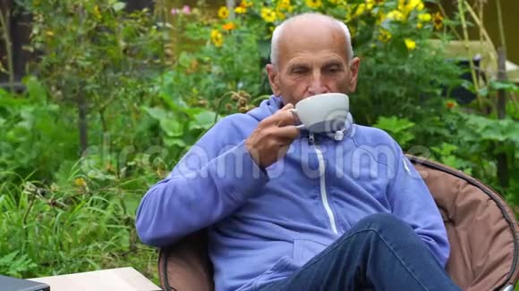 老人在盛开的花园里坐在舒适的扶手椅上喝茶视频的预览图