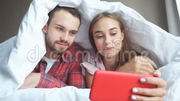 年轻夫妇躺在白色毯子下用手机看视频视频的预览图