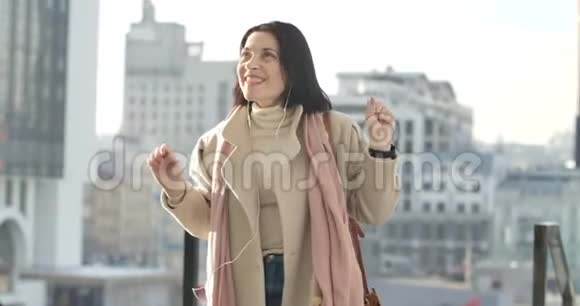 美丽的高加索女人戴着耳机在摩天大楼的背景下听音乐优雅的商务小姐视频的预览图