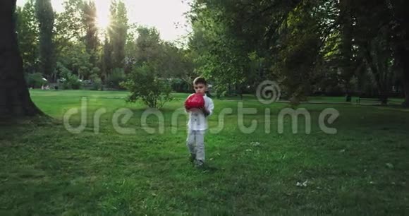 一个漂亮的小男孩穿着一个红色的球走在绿色的大公园中央的草地上视频的预览图