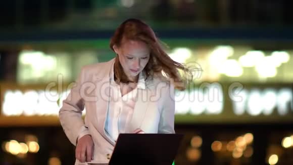 年轻漂亮的女性晚上坐在外面的笔记本电脑上使用智能手机视频的预览图