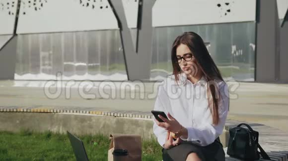 美丽的商务女性一边吃羊角面包一边在手机上聊天一边在户外微笑视频的预览图