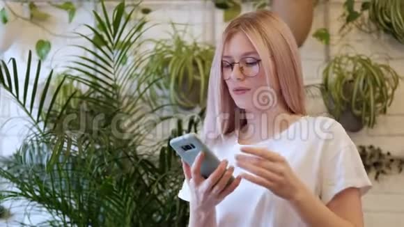 年轻漂亮的金发女郎戴着眼镜在咖啡馆的桌子上随着智能手机上的音乐跳舞视频的预览图