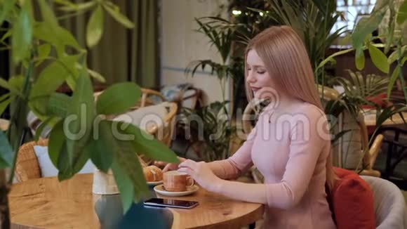 年轻漂亮的金发女人穿着粉红色的裙子坐在咖啡馆的桌子旁喝茶视频的预览图