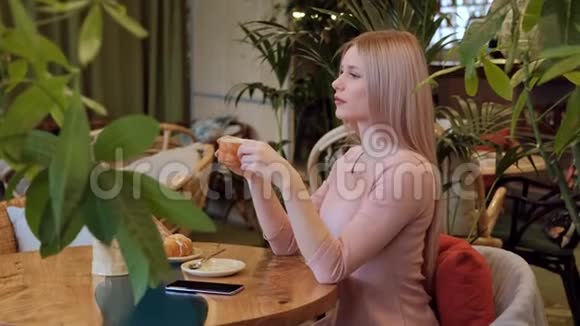 年轻漂亮的金发女人穿着粉红色的裙子坐在咖啡馆的桌子旁喝茶视频的预览图