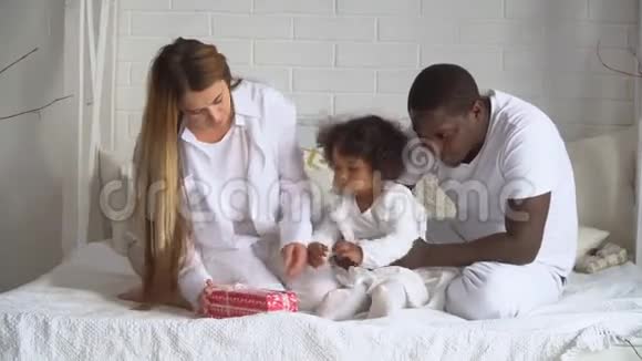 年轻幸福的多民族家庭的母亲非洲父亲和穿着白色衣服的小女儿正坐在床上视频的预览图