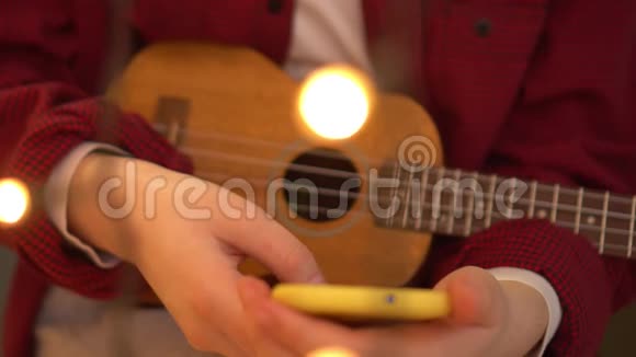 年轻的时髦女人弹着四弦琴吉他一个女人在智能手机上读一首歌背景灯在燃烧视频的预览图
