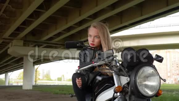 年轻女孩骑着皮夹克坐在城市背景下的停车摩托车上骑摩托车的摇滚女孩视频的预览图