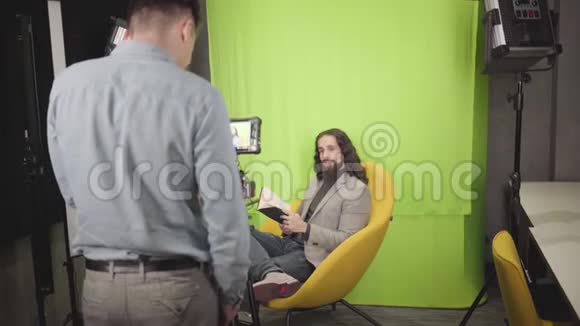 积极的中东男人坐在黄色的扶手椅上拿着书在镜头前交谈摄像机录像视频的预览图