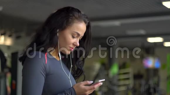 戴着耳机的年轻女孩在运动自行车上使用智能手机视频的预览图