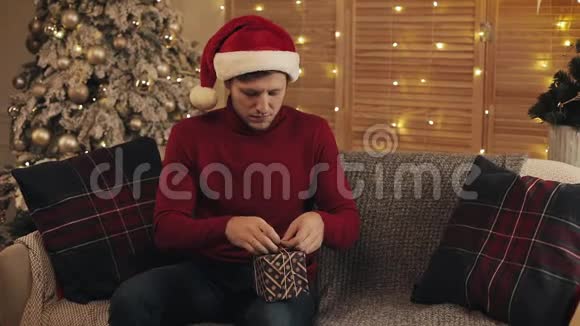 年轻的白种人戴着一顶桑塔帽坐在客厅圣诞树附近的沙发上在礼物上系着蝴蝶结视频的预览图