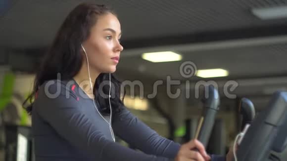 一个年轻漂亮的女孩戴着耳机坐在健身车上的肖像视频的预览图