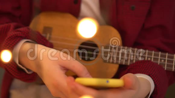 年轻的希普斯特女人演奏尤库莱吉他一个女人在智能手机上读一首歌背景灯在燃烧视频的预览图