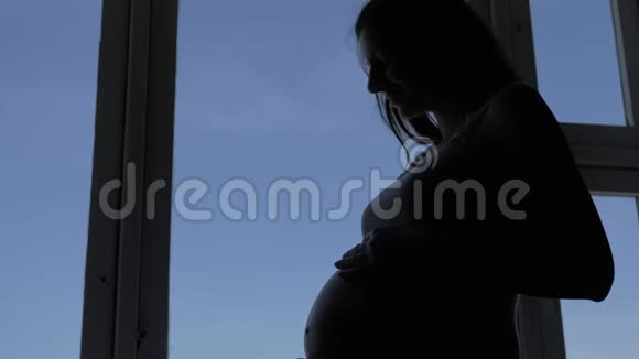 苗条孕妇踩着她的胃站在窗户附近的剪影视频的预览图
