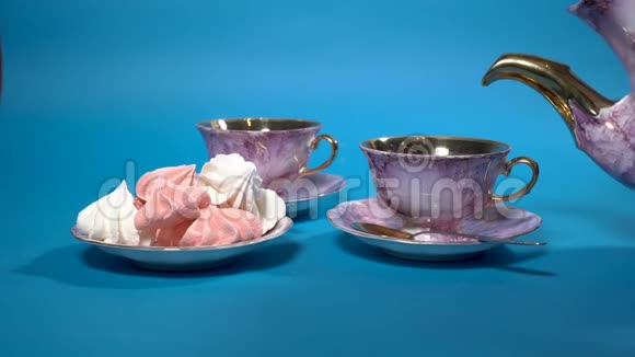 红茶被倒入蓝色背景上美丽的粉红色瓷杯中旁边有一碟棉花糖视频的预览图