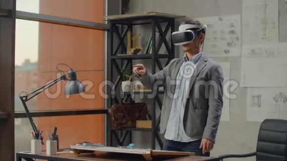 办公室里有大窗户的现代人建筑师站在虚拟现实头盔里用手势来管理视频的预览图