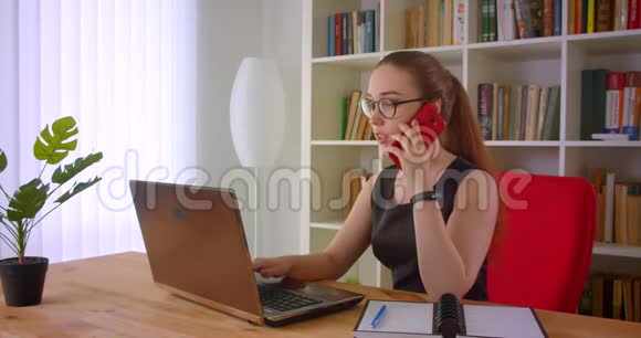 年轻漂亮的红发女商人戴着眼镜手提电脑在室内打电话视频的预览图