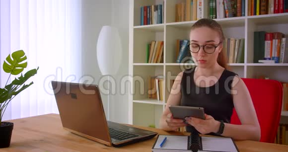年轻漂亮的红头发女商人戴着眼镜在办公室里用平板电脑坐在笔记本电脑前视频的预览图