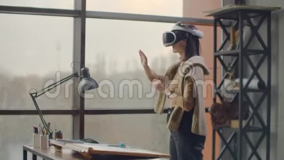 办公室里有大窗户的现代女建筑师站在虚拟现实头盔里用手势来管理视频的预览图
