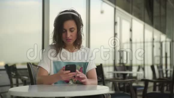 戴着耳机听音乐的年轻女子在户外咖啡馆用智能手机听音乐视频的预览图