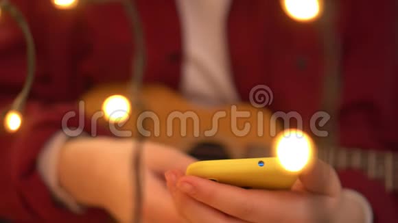 年轻的希普斯特女人演奏尤库莱吉他一个女人在智能手机上读一首歌背景灯在燃烧视频的预览图