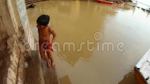 印度男孩穿着紧身衣在楼梯上凝视在肮脏的水附近用湿的材料编织洪水神圣的恒河木制的船视频的预览图