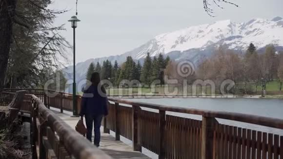 布鲁内特长头发的女孩背着背包走在木墩上背景是山湖后视图视频的预览图