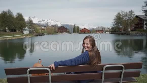 年轻女子坐在湖边的木凳上风景秀丽然后女孩回头微笑视频的预览图