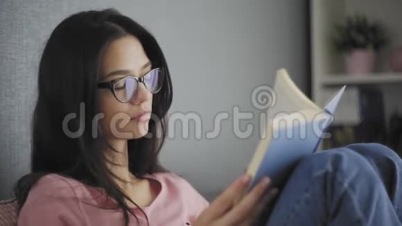 漂亮快乐的女学生戴着眼镜在一所现代化的房子的沙发上看书在找回到过去视频的预览图