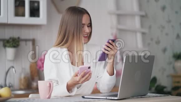 年轻女子坐在桌子上厨房里拿着笔记本电脑用两部手机视频的预览图