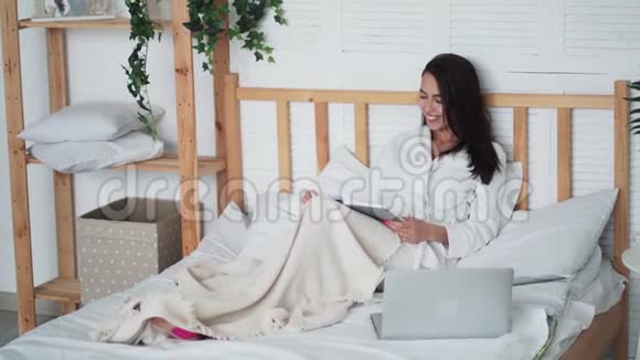 穿着白色浴袍的年轻美女躺在床上手提电脑看书视频的预览图