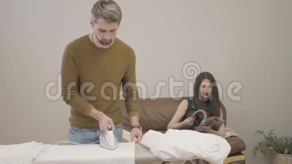 成年白种人男子熨烫深色头发的女人坐在后台丈夫在妻子休息时做家务视频的预览图