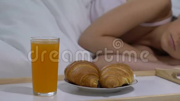 快乐的女人醒来看着床旁盘子里的牛角面包和果汁视频的预览图
