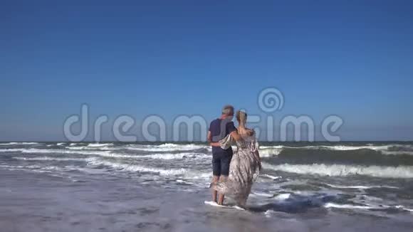 一个身材苗条的男人和一个穿着长裙的女人站在沙滩上迎风破浪视频的预览图