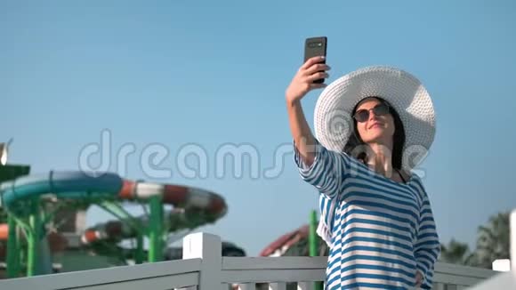 时尚旅游女性戴着墨镜和帽子在日落时分用智能手机自拍视频的预览图