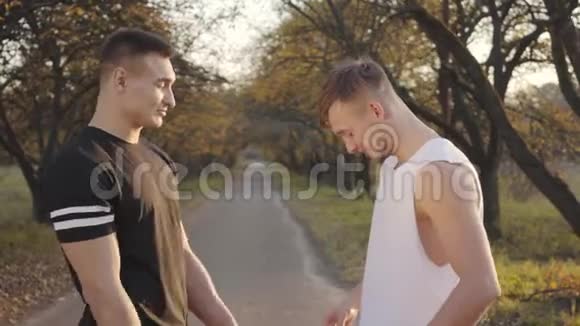 英俊的白人运动员站在秋天公园的阳光下炫耀他们的肌肉和笑声两个年轻人视频的预览图