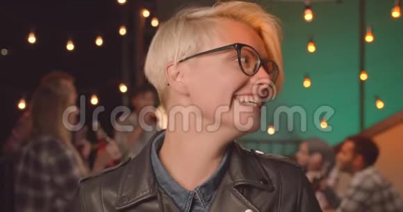 年轻漂亮的金发女性戴着眼镜看着镜头在舒适的聚会上愉快地笑着视频的预览图