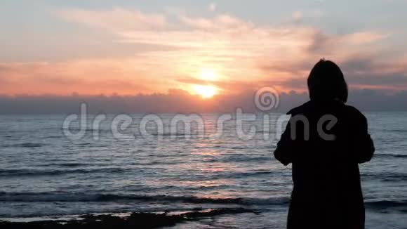 年轻女孩看日落的剪影可爱的女孩穿着休闲装站在岩石海岸看着海洋西尔胡特视频的预览图