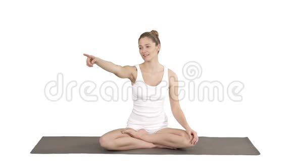 年轻迷人的女人坐在莲花的姿势指着什么或某人而说话的白色背景视频的预览图