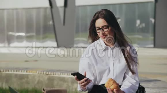 一位成功的公司女性一边在手机上聊天一边在户外微笑一边吃羊角面包视频的预览图