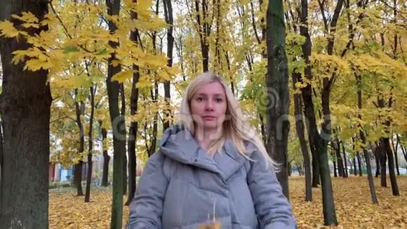 一个穿着温暖衣服的金发女郎站在秋天的公园里扔了一堆枫叶视频的预览图