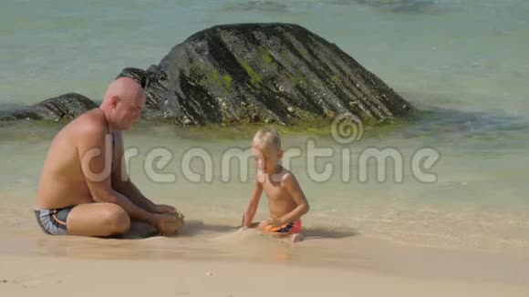 父亲向躺在海水里的滑稽儿子展示了潮湿的沙子视频的预览图