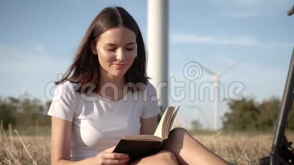 在风力发电站背景下一位坐在电动滑板车旁边的迷人女孩的肖像视频的预览图
