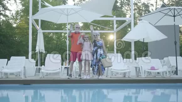 一对幸福的成熟夫妇和一个小孙女站在游泳池旁对着镜头挥手视频的预览图