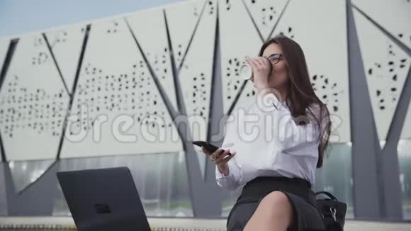 漂亮的商务女性戴着眼镜喝着咖啡看着她的智能手机视频的预览图