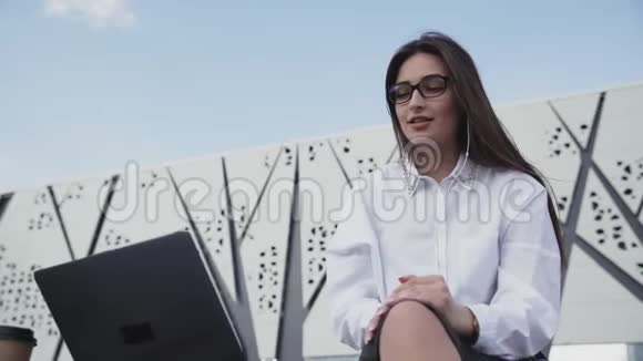 年轻的商务女性带着耳机在Skype上聊天使用笔记本电脑坐在户外视频的预览图
